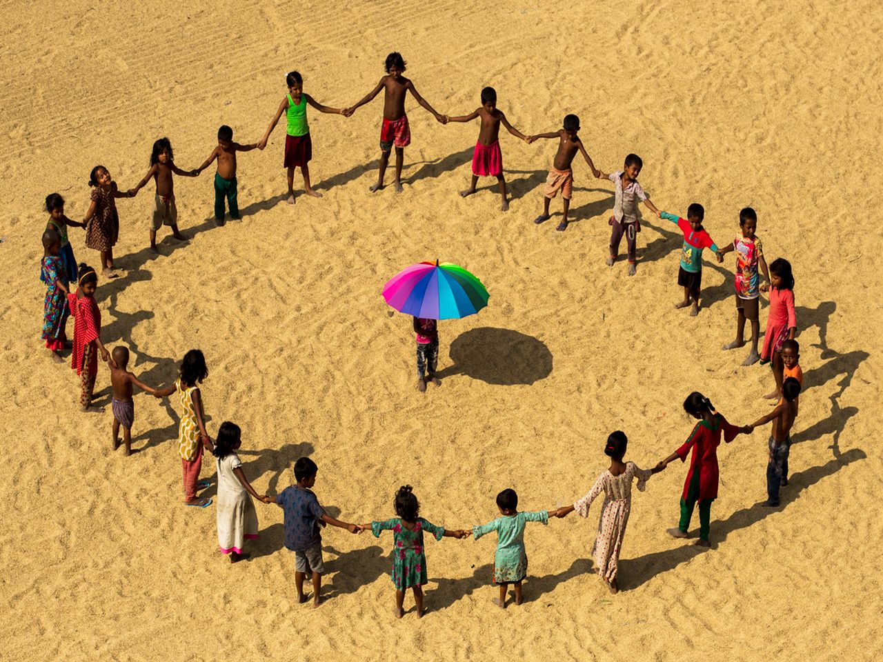 Aerial view of children holding hands in a circle on sandy ground with colorful umbrella in Chittagong, Bangladesh.